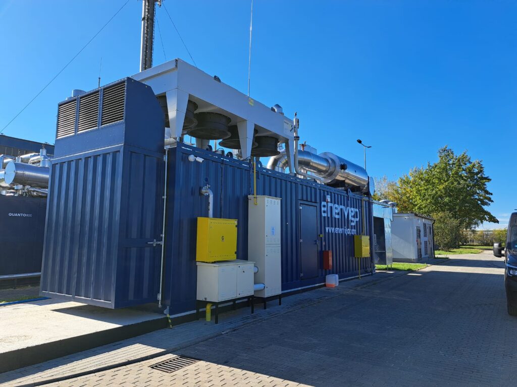Ein großer blauer Energiepartner-Industriecontainer mit Rohren und Entlüftungsöffnungen steht im Freien auf einer gepflasterten Fläche unter einem klaren blauen Himmel. Im Hintergrund ist ein Baum zu sehen.