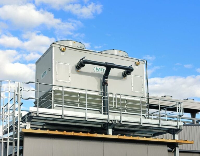 Ein großes industrielles Kühlaggregat mit zwei Ventilatoren, das innovative Energielösungen unterstützt, ist auf einer Plattform mit Metallgeländer vor einem blauen Himmel montiert. Das Gerät ist mit "MITA" beschriftet und durch schwarze Rohre verbunden.