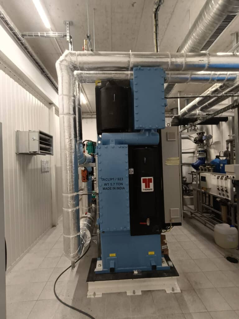 Eine große Industriemaschine mit der Aufschrift "TACLYP/123 MPT/100A Made in India" steht in einem sauberen, gefliesten Raum und zeigt die moderne Industrielle Energieversorgung inmitten von Metallrohren, Kanälen und anderen Energieanlagen im Hintergrund.