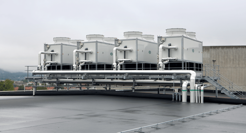 Vier industrielle Kühltürme mit miteinander verbundenen Rohren, die zu fortschrittlichen Energielösungen gehören, sind auf dem flachen Dach eines großen Gebäudes installiert, wobei im Hintergrund ein bewölkter Himmel und weit entfernte Bäume zu sehen sind.