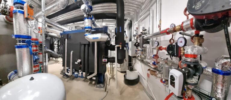 Ein Maschinenraum mit verschiedenen isolierten Rohren, Ventilen, Manometern und einem großen schwarzen Industrieaggregat; silberne Isolierung und blaue und rote Armaturen sind in diesem gut beleuchteten Raum für moderne Industrielle Energieversorgung sichtbar.