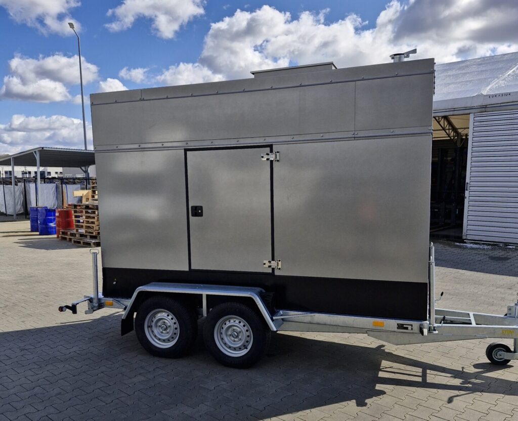 Ein großer, metallischer, rechteckiger Anhänger für mobile Energieerzeugung steht auf einer gepflasterten Fläche unter einem teilweise bewölkten Himmel. Mit zwei Rädern und einer Tür ist dieser Anhänger an einer Anhängerkupplung befestigt und bereit für innovative Energielösungen für unterwegs.