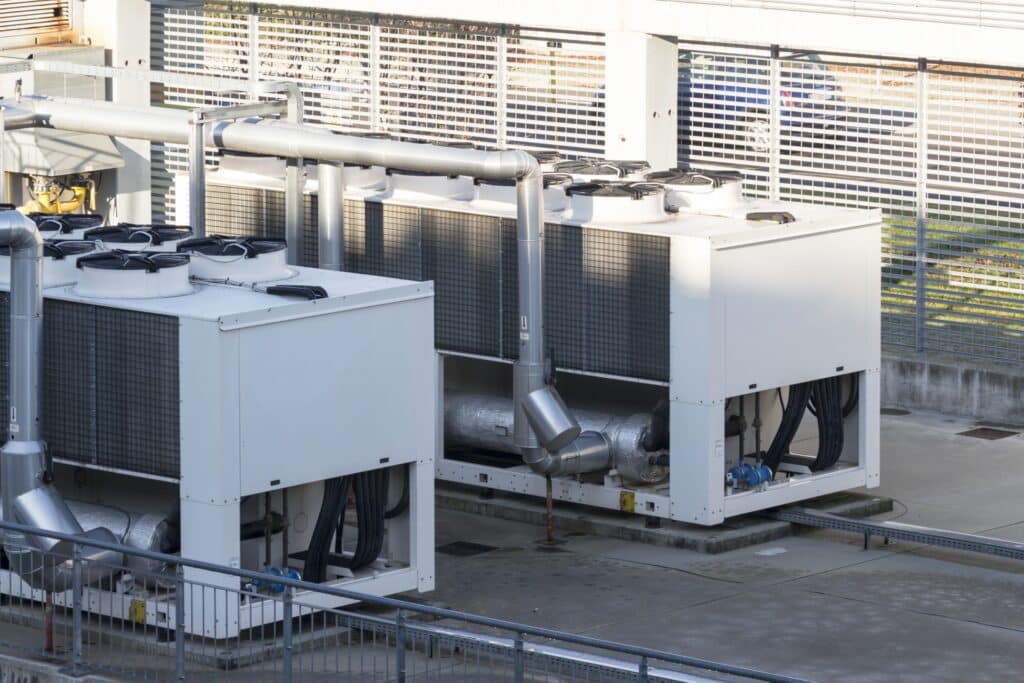 Zwei große, für die moderne Energieerzeugung unverzichtbare Industrieklimageräte mit sichtbaren Lüftungsrohren und -gittern sind im Freien auf einer Betonfläche installiert, die von Metallzäunen und Geländern umgeben ist.
