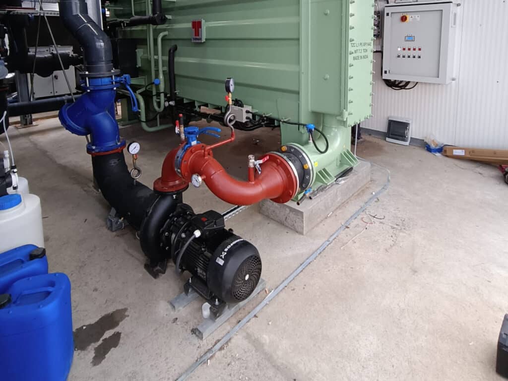Industrielles Pumpensystem mit schwarzen Rohren, einem roten Ventil, Messgeräten, einer grünen Maschine und einer elektrischen Schalttafel für moderne Energieanlagen. Auf dem Betonboden sind auch blaue Behälter und andere Geräte zu sehen.