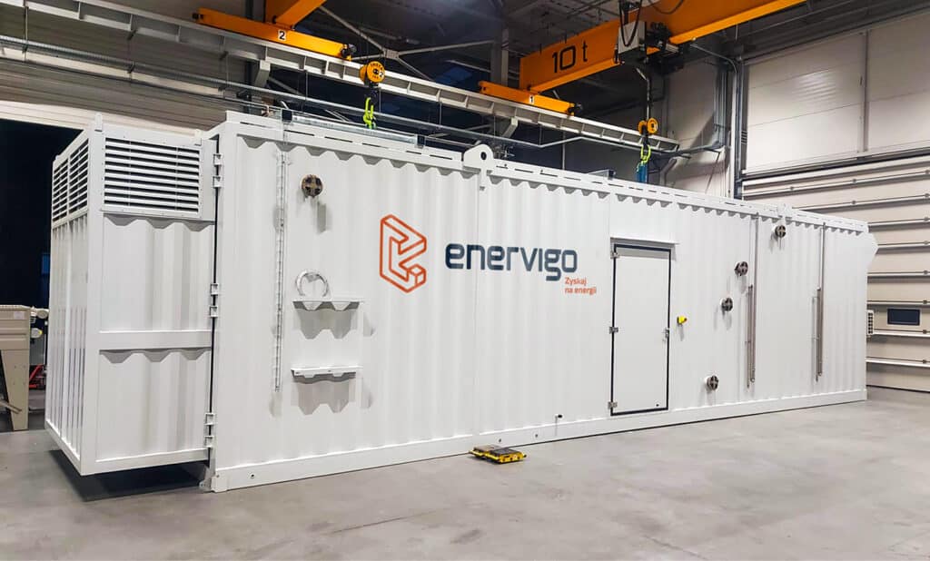 Ein großer weißer Industriecontainer mit der Aufschrift "enervigo" steht in einem Lagerhaus oder einer Fabrik unter einem gelben Brückenkran und präsentiert innovative Energielösungen mit Lüftungsöffnungen, Metallvorrichtungen und einer Tür an der Außenseite.