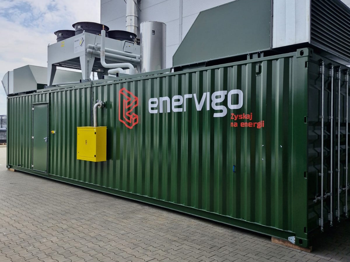 Ein großer Ökostrom-Container mit der Aufschrift "Enervigo", einem Logo und dem Schriftzug "Zyskaj na energii" steht auf einer gepflasterten Fläche neben einem Gebäude, das die industrielle Energieversorgung unterstützt und auf dem fortschrittliche Geräte montiert sind.