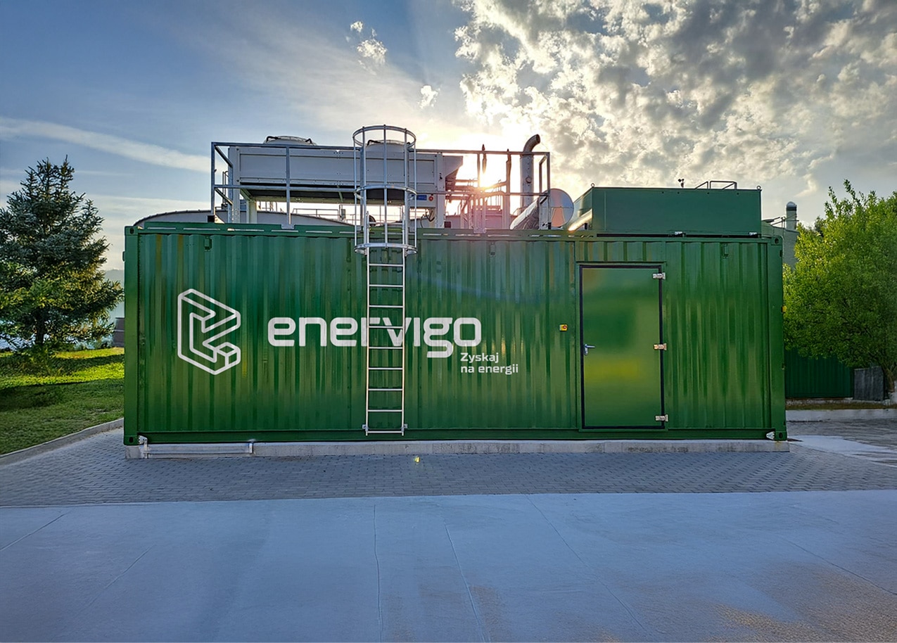 Eine große grüne Containeranlage mit dem "Enervigo"-Logo und polnischem Text steht unter freiem Himmel auf einer gepflasterten Fläche, umgeben von Bäumen, unter teilweise bewölktem Himmel - und zeigt moderne Energieanlagen für die industrielle Energieversorgung.