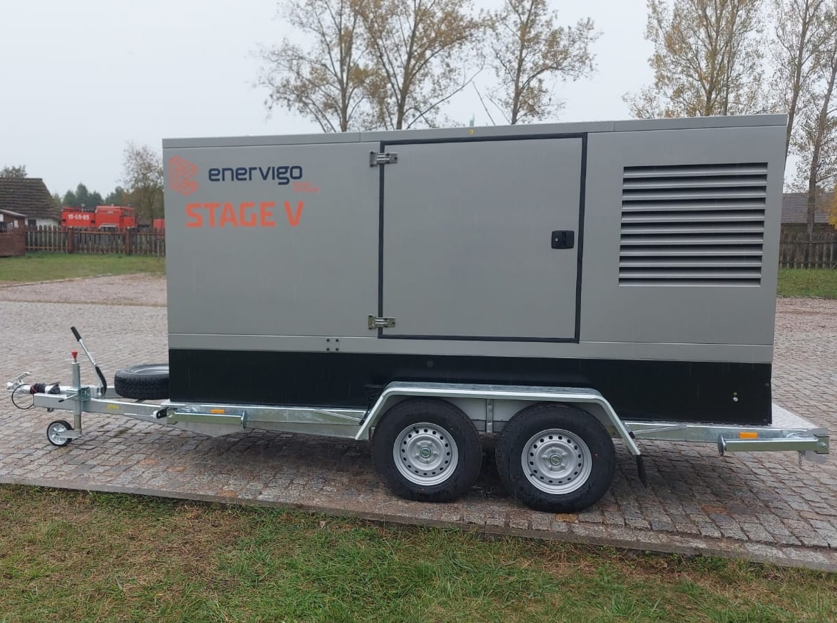 Ein mobiles Stromaggregat mit der Aufschrift "Enervigo STAGE V" ist auf einem zweiachsigen Anhänger montiert, der an einem bewölkten Tag zwischen Bäumen und Gebäuden auf Kopfsteinpflaster geparkt ist und die innovative Energieerzeugung für die industrielle Energieversorgung präsentiert.