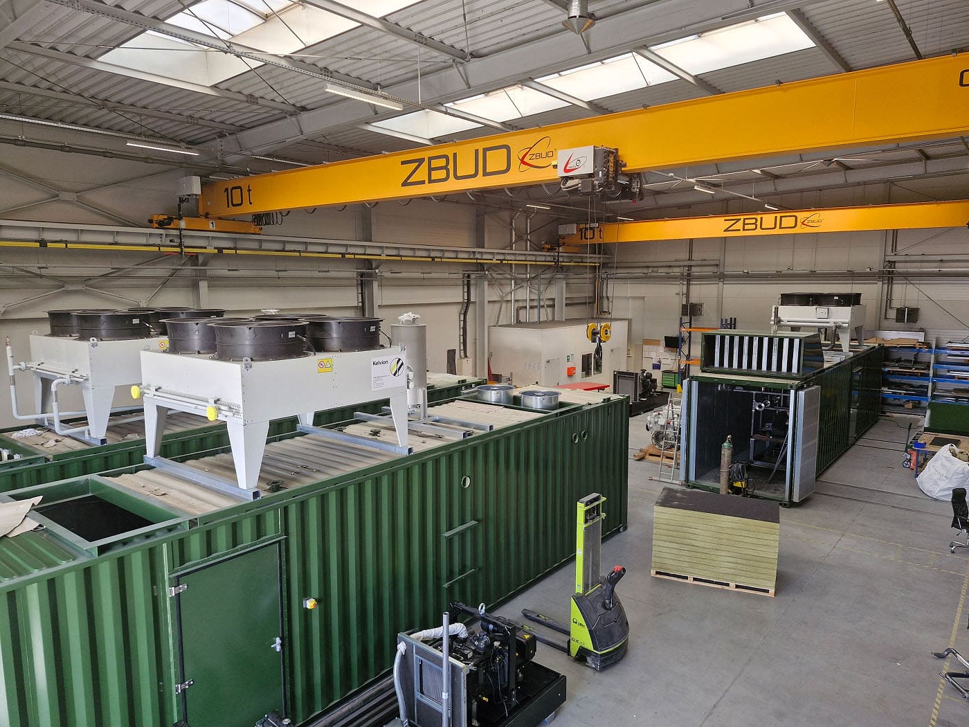 Das Innere eines großen Industrielagers mit grünen Schiffscontainern, Maschinen, Kühlaggregaten auf den Containern, gelben "ZBUD"-Kränen und verschiedenen Werkzeugen, die die innovative Industrielle Energieversorgung und fortschrittliche Energielösungen zeigen.
