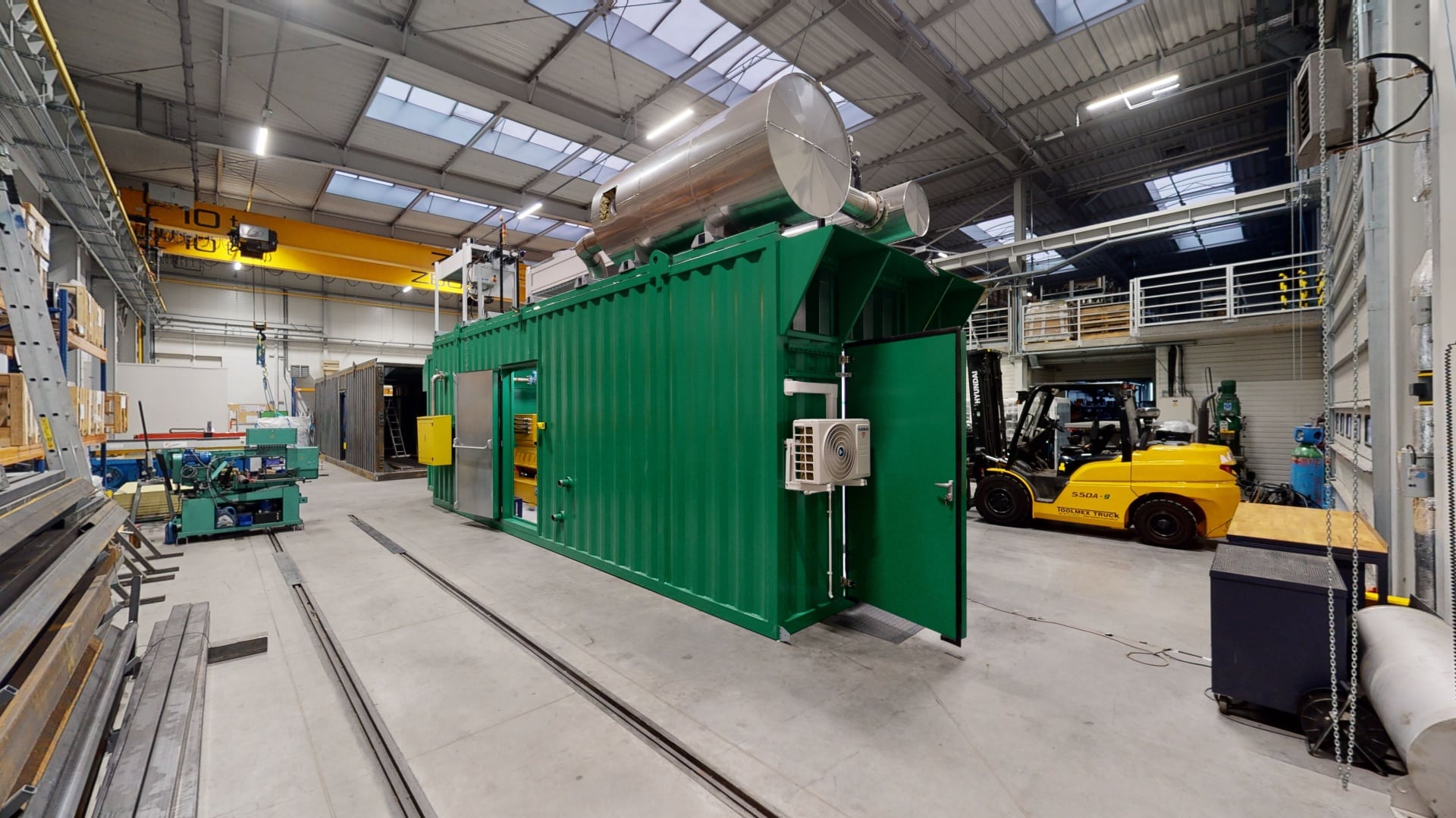 Ein großer grüner Industriecontainer für Energieerzeugung mit Metallrohren auf der Oberseite befindet sich in einer geräumigen Lagerhalle mit hohen Decken, Oberlichtern und verschiedenen Geräten, darunter ein gelber Gabelstapler in der Nähe.