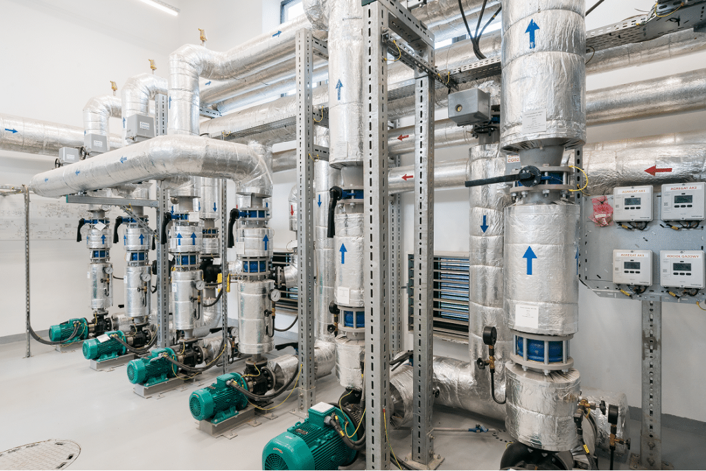 Ein Maschinenraum mit mehreren isolierten Rohren, Ventilen, Messgeräten und grünen Industriepumpen demonstriert effiziente Energieerzeugung; blaue Pfeile zeigen die Flussrichtung an. Die Energieanlagen sind in einem sauberen, gut beleuchteten Raum übersichtlich angeordnet.
