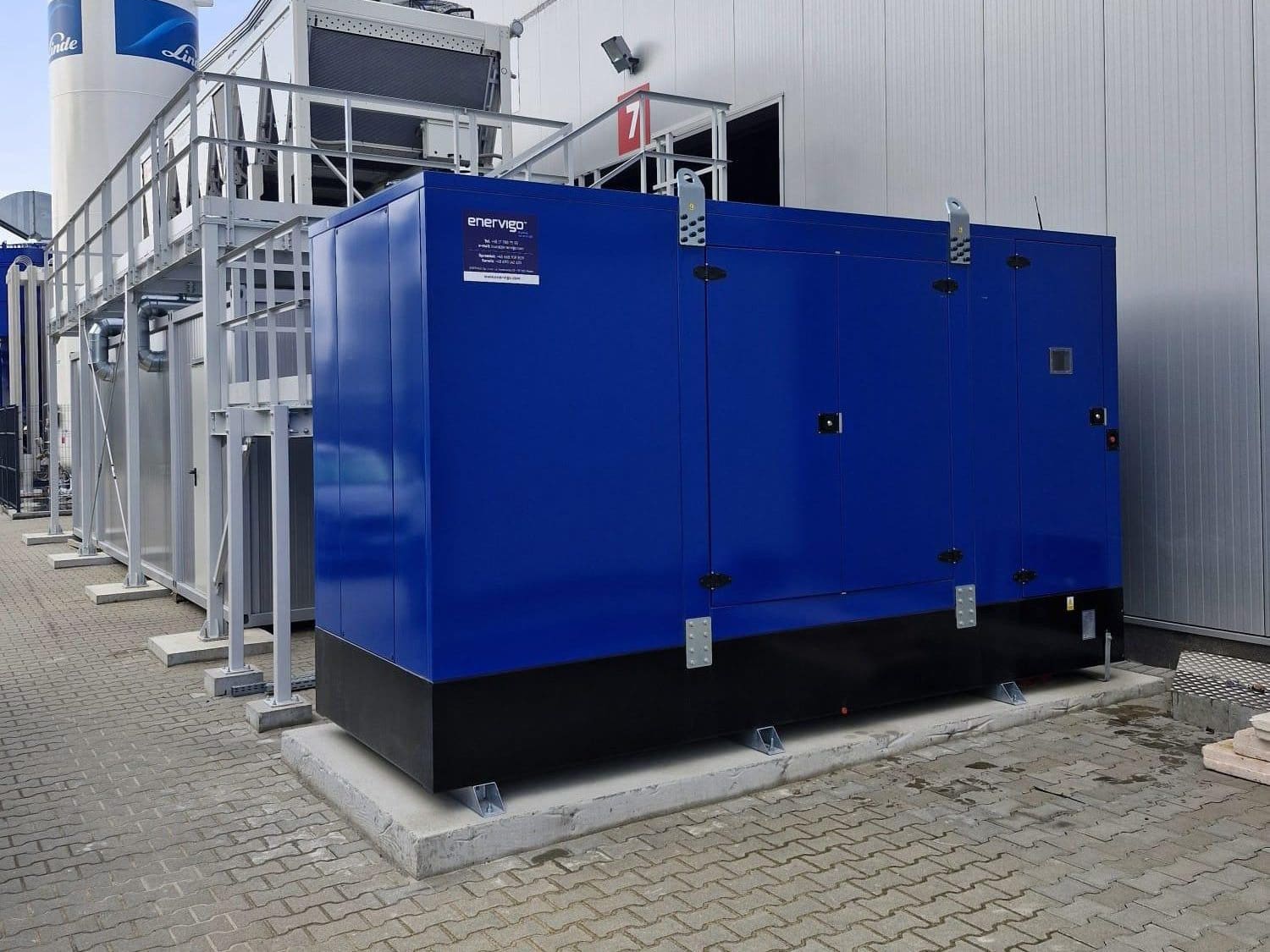 Ein großer blauer Industriegenerator, der zu modernen Energieanlagen gehört, ist auf einer Betonplattform vor einem Industriegebäude installiert; im Hintergrund sind Metallstrukturen und -geräte zu sehen.