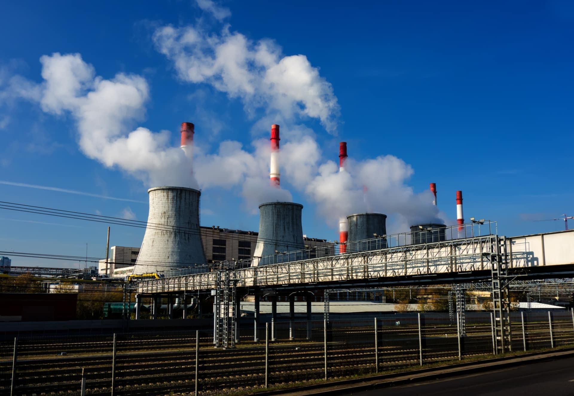 Ein Kraftwerk für Energieerzeugung mit mehreren großen Kühltürmen und rot gestreiften Schornsteinen stößt unter blauem Himmel Dampf und Rauch aus, im Vordergrund sind Eisenbahnschienen und Metallkonstruktionen zu sehen.