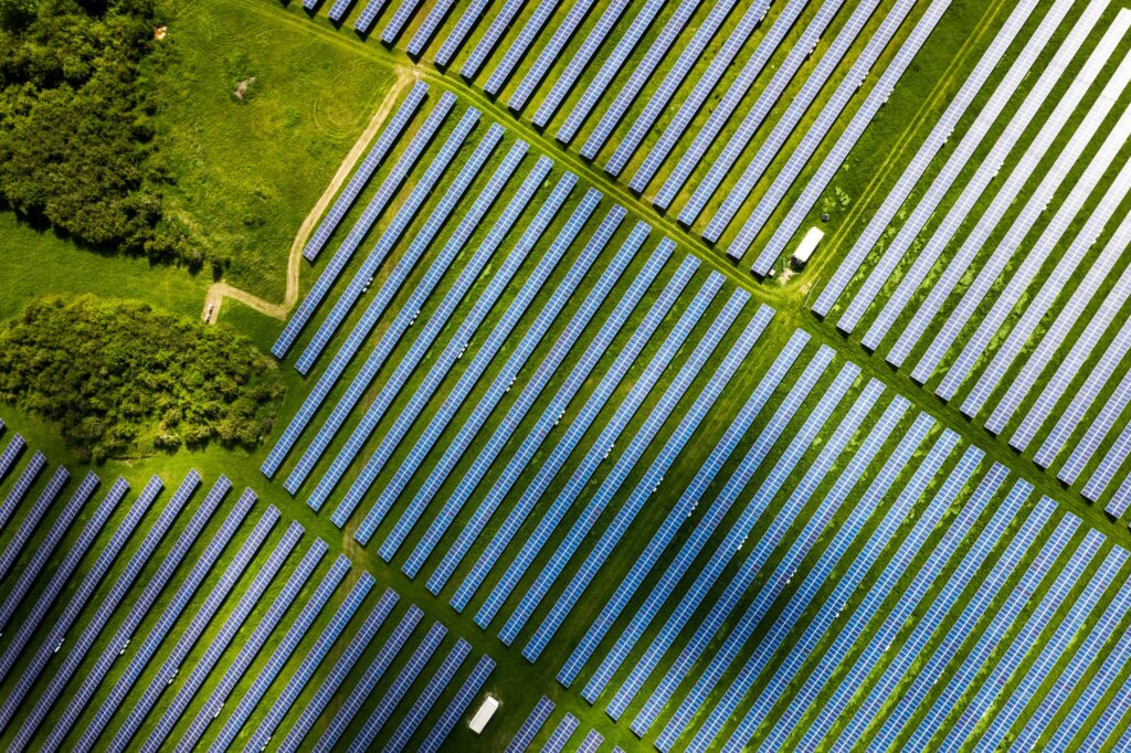 Luftaufnahme eines großen Solarparks - Reihen von blauen Paneelen der innovativen Energieanlagen der Industriellen Energieversorgung, die sich über grüne Felder mit Bäumen erstrecken und teilweise von vorbeiziehenden Wolken beschattet werden.