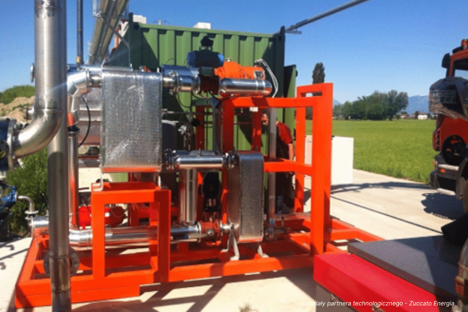 Industrielle Außenanlagen für moderne Energielösungen mit glänzenden Metallrohren und Bauteilen auf einem orangefarbenen Rahmen, aufgestellt auf einer Betonplattform in der Nähe einer Wiese und von Bäumen unter einem sonnigen Himmel.