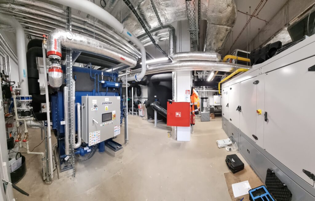 Ein großer Industrieraum, der für eine effiziente Energieerzeugung konzipiert wurde, weist komplexe Maschinen, Schalttafeln, Metallrohre und Lüftungskanäle auf. Ein roter Notfallkasten und verschiedene Werkzeuge sind in diesem sauberen, organisierten Raum mit Betonböden und Sichtdecken zu sehen.